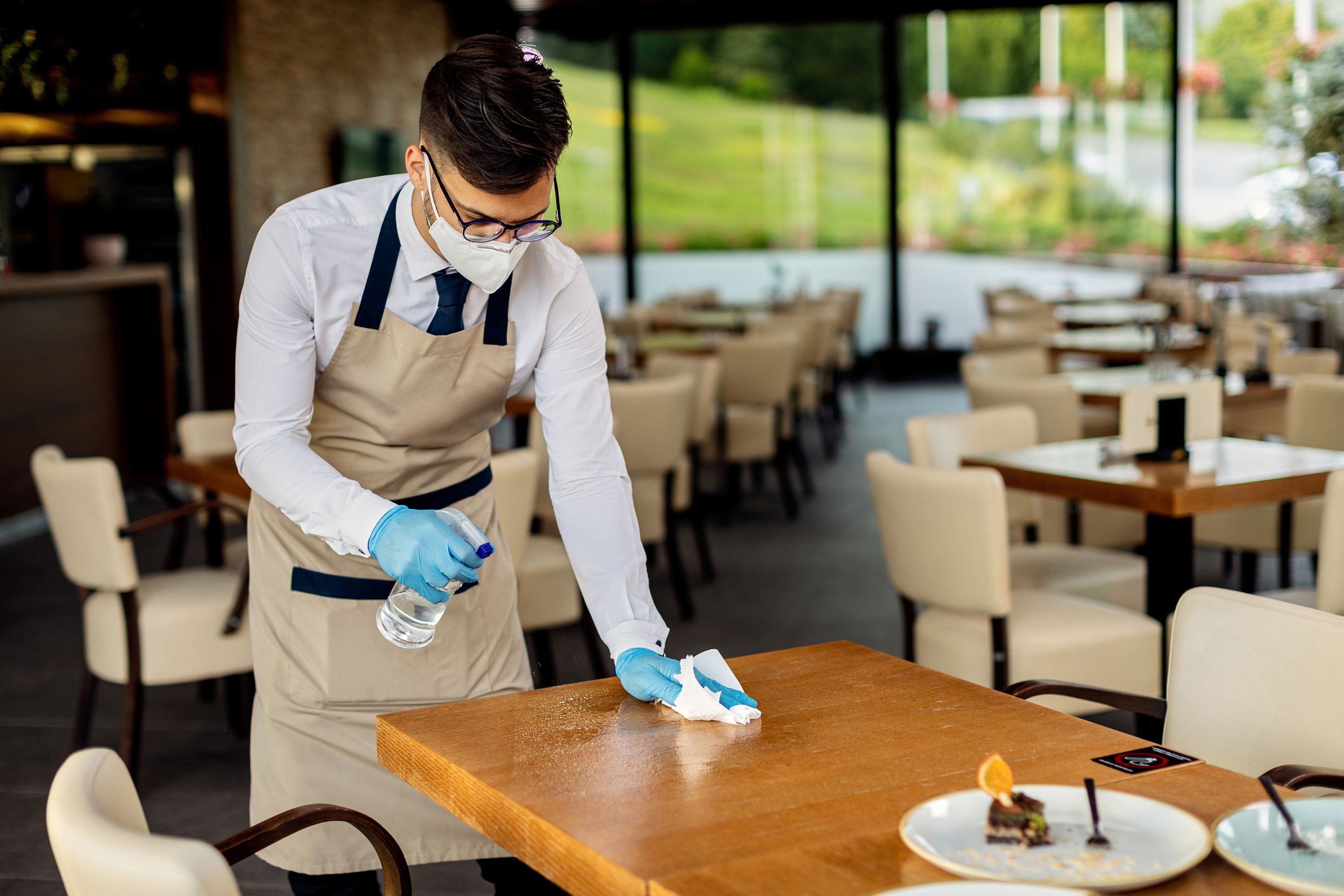 Waiter with protective face mask disinfecting tables in a cafe d